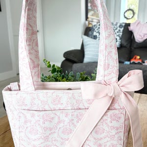 May include: A quilted fabric tote bag featuring a white base with a pink floral toile pattern. The bag includes a single wide shoulder strap, a front exterior pocket, and a solid light pink fabric bow attached to the side of the bag opening.