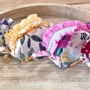 May include: Four small, floral-patterned zippered pouches with ruffled edges. The pouches feature various colors, including purple, yellow, pink, and red, and are adorned with gold-colored hardware and small wrist straps. The pouches are arranged on a wooden tray.