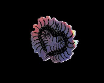 Puede incluir: Una pegatina holográfica con un diseño de ciempiés negro en forma de corazón. La pegatina tiene un borde festoneado y un degradado de color rosa, morado y amarillo. El fondo es negro.