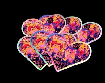 Puede incluir: Una colección de pegatinas en forma de corazón con un acabado holográfico. Cada pegatina presenta una ilustración de un gato dentro de un marco en forma de corazón, rodeada de imágenes y texto adicionales. Las pegatinas tienen una vibrante paleta de colores rosa, morado y amarillo.