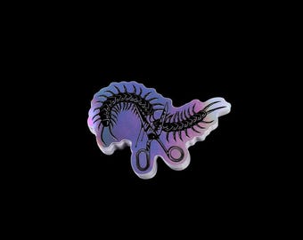 Puede incluir: Pin holográfico con un esqueleto de ciempiés estilizado con tijeras. El pin tiene un degradado de tonos morados, azules y rosas. El diseño está sobre un fondo negro.