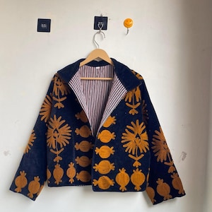 以下が含まれることがあります： ネイビーブルーのジャケット。金色の花の刺繍が施されています。裏地はストライプで、クラシックな襟が付いています。デザインには、様式化された花柄と円形のパターンが含まれており、ユニークな外観を作り出しています。木製のハンガーに掛けられています。