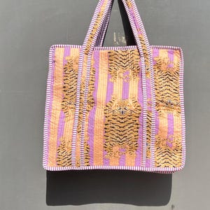 Op de afbeelding: Een grote tote bag met een paars en oranje gestreept patroon en een tijgerprint. De tas heeft een brede bovenopening en twee handvatten.