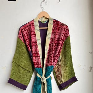 May include: A patchwork robe with a tie belt, displayed against a white wall. The robe is a vibrant mix of colours and patterns, including pink, green, blue, and cream. The sleeves are green with a purple trim, and the body has a mix of patterned fabrics.