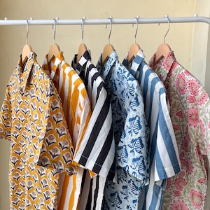 May include: A collection of short-sleeved shirts hanging on a white rack. The shirts feature various patterns, including floral, stripes, and geometric designs. Colors include yellow, black, white, blue, and pink. The shirts are made of a lightweight fabric.