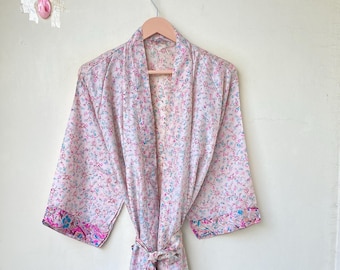 May include: A light pink floral patterned robe with a matching belt, hanging on a wooden hanger. The robe has blue and pink accents on the sleeves and trim. The robe is made of a lightweight fabric.