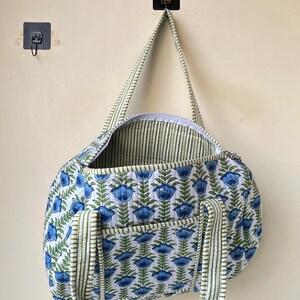 May include: A quilted duffel bag with a blue floral pattern on a white background. The bag has green and white striped handles and trim. The bag is hanging on a wall.