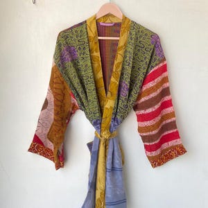 May include: A colorful, patterned robe with a tie belt, hanging on a wooden hanger. The robe features a mix of green, purple, red, and yellow fabrics. The sleeves have striped and patterned sections.