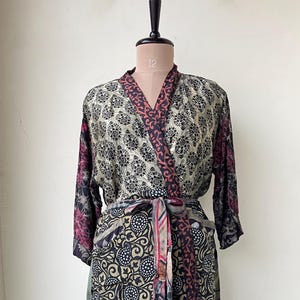 May include: A patterned robe with a floral design in black, gold, and burgundy. The robe is tied at the waist with a matching belt. The sleeves are long, and the robe is displayed on a mannequin. The robe is made of a silky material.