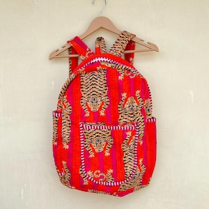 Puede incluir: Una mochila roja y rosa con un diseño de estampado de tigre. La mochila tiene un bolsillo frontal y correas ajustables. El estampado de tigre presenta tigres naranjas y negros sobre un fondo a rayas rojas y rosas. La mochila está colgada de una percha de madera.