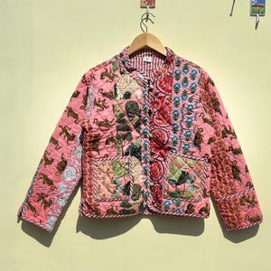 May include: A quilted jacket with a patchwork design. The jacket features a pink base color with various patterns, including floral and animal motifs. It has a round collar, long sleeves, and two front pockets. The jacket is hanging on a wooden hanger.