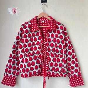 May include: A red and white quilted jacket featuring a strawberry print. The jacket has a red gingham collar, cuffs, and tie closure. It is displayed on a wooden hanger.