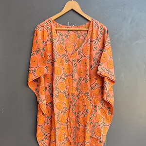 May include: A long, peach-colored kaftan with a floral print featuring orange flowers and green leaves. The kaftan has a V-neck and short sleeves.