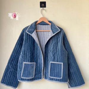 Könnte beinhalten: Blau gesteppte Jacke mit Samtstruktur, mit weiß-blau gestreiftem Futter und Besatz. Die Jacke hat zwei Vordertaschen und hängt an einem Holzbügel.