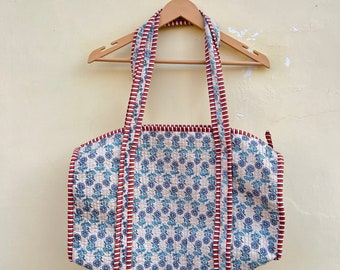 May include: A white and blue patterned tote bag with red and white trim. The bag has two straps and is hanging on a wooden hanger.