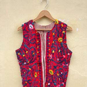 Puede incluir: Un chaleco rojo sin mangas con un patrón de bordado floral. El chaleco tiene una cremallera y un forro a rayas.