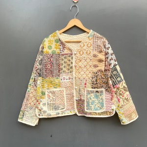 May include: A cream-colored quilted jacket with a patchwork design featuring various floral and geometric patterns in shades of pink, red, yellow, blue, and green. The jacket has a button-up front and two patch pockets.