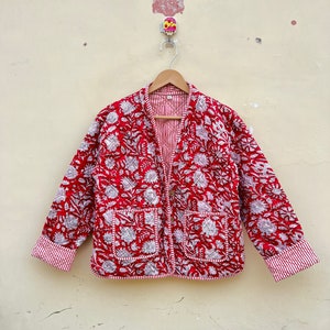 Puede incluir: Una chaqueta acolchada roja y blanca con estampado floral. La chaqueta tiene cierre de botón y dos bolsillos.