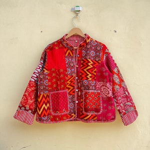Op de afbeelding: Een rood gewatteerd jack met een patchwork-ontwerp. Het jack heeft een mandarijnsluiting, lange mouwen en een knoopsluiting. De stof is een mix van verschillende patronen en kleuren, waaronder rood, oranje, geel, blauw en groen.