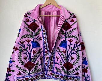 以下が含まれることがあります： ピンク色のベルベットジャケット、花の刺繍入り。 ジャケットは襟、長袖、ストライプの裏地付き。 刺繍には赤、青、緑の花柄が含まれています。 ジャケットは木製のハンガーに掛けられています。