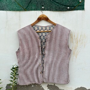 May include: A sleeveless vest with a striped pattern in white and dark brown. The vest has a button-up front and a floral patterned lining. It hangs on a wooden hanger against a white wall.