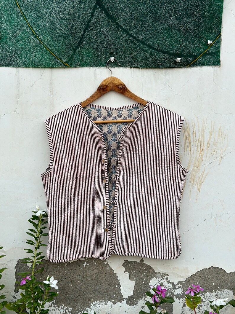 May include: A sleeveless vest with a striped pattern in white and dark brown. The vest has a button-up front and a floral patterned lining. It hangs on a wooden hanger against a white wall.