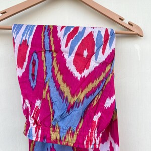 May include: Pink, blue, and gold patterned pants with an elastic waistband. The pants are hanging on a wooden hanger.