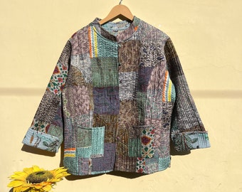 May include: A patchwork quilted jacket with a mandarin collar and button closure. The jacket is made of various fabrics in shades of brown, green, blue, and purple. The jacket has a pocket on the left side and is hanging on a wooden hanger.