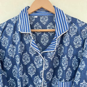 K&ouml;nnte beinhalten: Ein blaues und wei&szlig; gestreiftes Baumwoll-Pyjama-Oberteil mit wei&szlig;em Blumenmuster und wei&szlig;em Besatz. Das Oberteil hat einen Knopfleisten vorne und eine Tasche auf der linken Seite. Das Gr&ouml;&szlig;enetikett zeigt "XL".