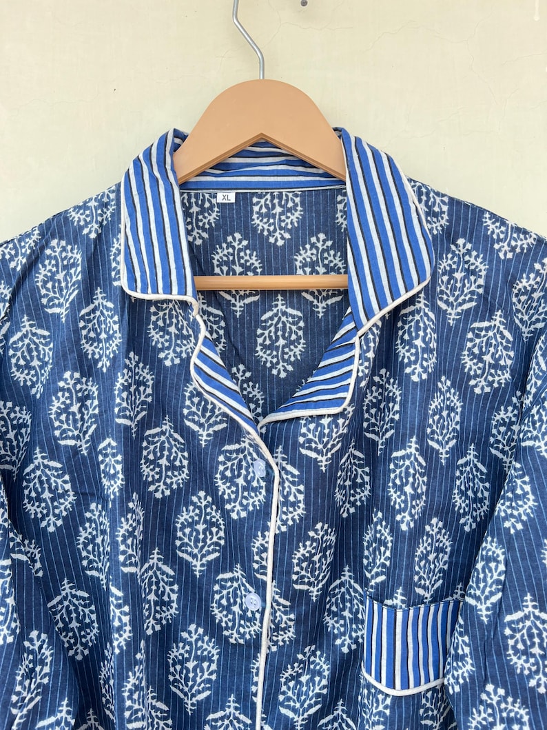 K&ouml;nnte beinhalten: Ein blaues und wei&szlig; gestreiftes Baumwoll-Pyjama-Oberteil mit wei&szlig;em Blumenmuster und wei&szlig;em Besatz. Das Oberteil hat einen Knopfleisten vorne und eine Tasche auf der linken Seite. Das Gr&ouml;&szlig;enetikett zeigt "XL".