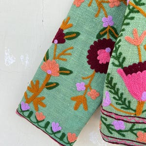 May include: A green jacket with colorful floral embroidery. The jacket has a long sleeve and a  pink, orange, and purple floral pattern.