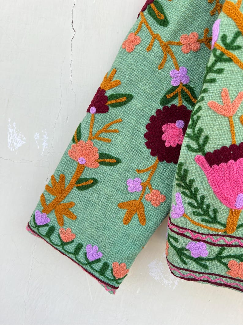 May include: A green jacket with colorful floral embroidery. The jacket has a long sleeve and a  pink, orange, and purple floral pattern.