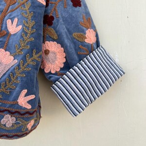 May include: A blue velvet jacket with embroidered floral details. The jacket has a striped cuff detail.