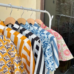 May include: A collection of patterned shirts hanging on a white rack. The shirts feature various designs, including yellow floral, orange and white stripes, black and white geometric, blue floral, blue and white stripes, and pink floral patterns.