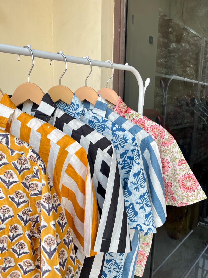 May include: A collection of patterned shirts hanging on a white rack. The shirts feature various designs, including yellow floral, orange and white stripes, black and white geometric, blue floral, blue and white stripes, and pink floral patterns.