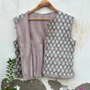 May include: Reversible quilted vest with a floral pattern on one side and black and white stripes on the other. The vest is sleeveless and has wooden button closures. The floral side features blue and gray flowers on a beige background.