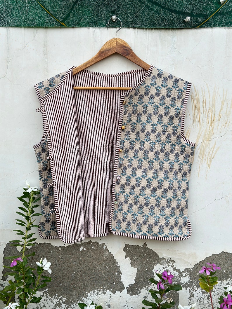 May include: Reversible quilted vest with a floral pattern on one side and black and white stripes on the other. The vest is sleeveless and has wooden button closures. The floral side features blue and gray flowers on a beige background.