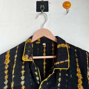 Może przedstawiać: Czarna koszula z ż&oacute;łto-pomarańczowym wzorem tie-dye. Koszula ma ż&oacute;łte lam&oacute;wki na kołnierzu, mankietach i kieszeni. Wisi na r&oacute;żowym, drewnianym wieszaku.