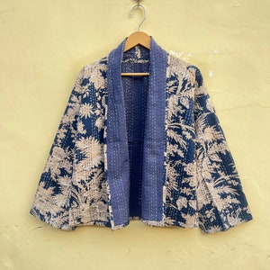 May include: A blue and white floral patterned quilted jacket with a long sleeve and a wide collar. The jacket is hanging on a wooden hanger against a yellow wall.