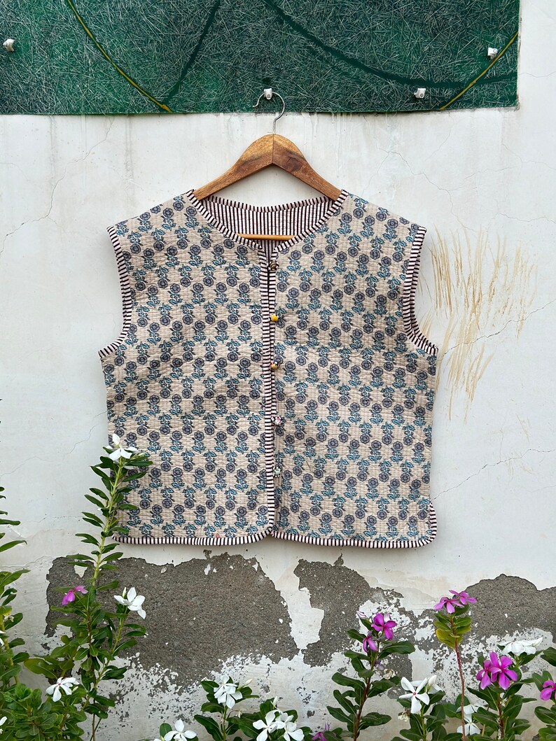 May include: A reversible, sleeveless vest with a floral pattern in blue and beige. The vest has a striped trim and is buttoned with gold-colored buttons. It hangs on a wooden hanger against a white wall.