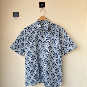 May include: A short-sleeved button-down shirt with a blue and white leaf pattern. The shirt is made of a lightweight fabric and features a classic collar and wooden buttons. The shirt is hanging on a wooden hanger.