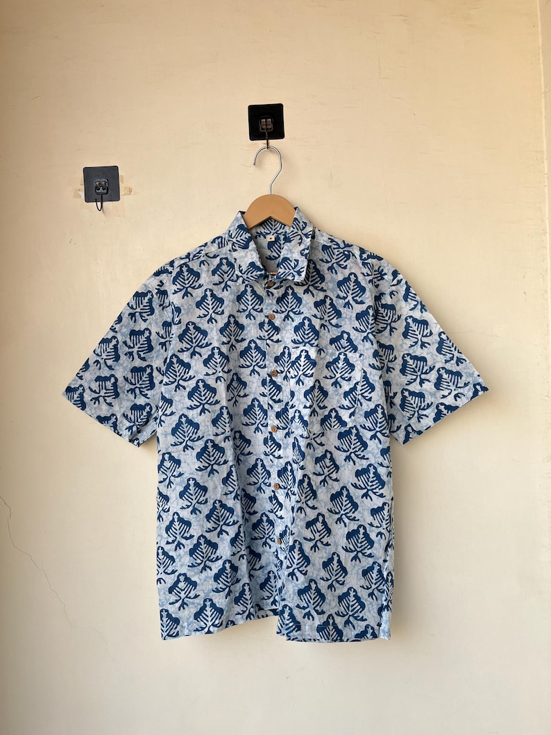 May include: A short-sleeved button-down shirt with a blue and white leaf pattern. The shirt is made of a lightweight fabric and features a classic collar and wooden buttons. The shirt is hanging on a wooden hanger.