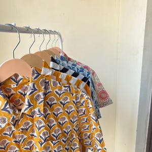 May include: A row of button-down shirts with floral patterns hanging on wooden hangers. The shirts feature various colors and designs, including yellow with a white and navy floral print, and blue with a white floral print. The shirts are made of a lightweight fabric.