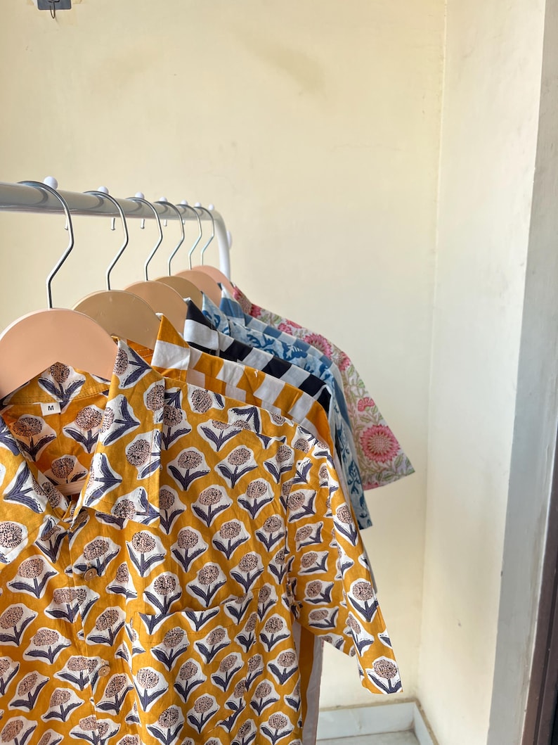 May include: A row of button-down shirts with floral patterns hanging on wooden hangers. The shirts feature various colors and designs, including yellow with a white and navy floral print, and blue with a white floral print. The shirts are made of a lightweight fabric.
