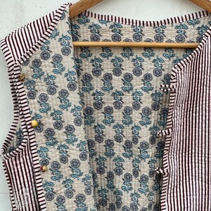 May include: A sleeveless quilted vest with a striped exterior in white and burgundy. The interior features a floral pattern in blue and gray on a cream background. The vest is fastened with small, decorative buttons.