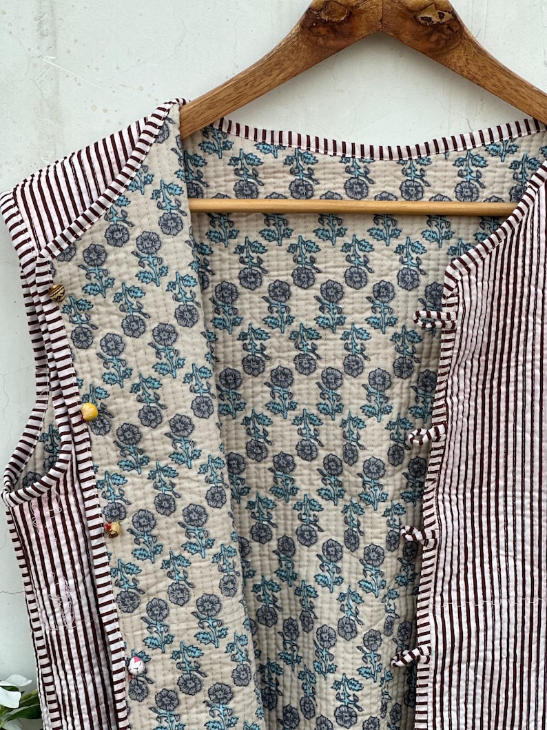 May include: A sleeveless quilted vest with a striped exterior in white and burgundy. The interior features a floral pattern in blue and gray on a cream background. The vest is fastened with small, decorative buttons.
