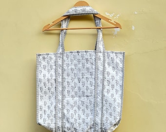 Op de afbeelding: Een witte tote bag met een zwart-witte bloemenprint die aan een houten hanger hangt. De tas heeft een zwarte bies en een zwart-wit gestreepte handvat.