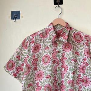 May include: Short-sleeved button-down shirt with a floral pattern. The shirt features a white background with a repeating pattern of pink flowers and green leaves. The shirt is hanging on a wooden hanger.