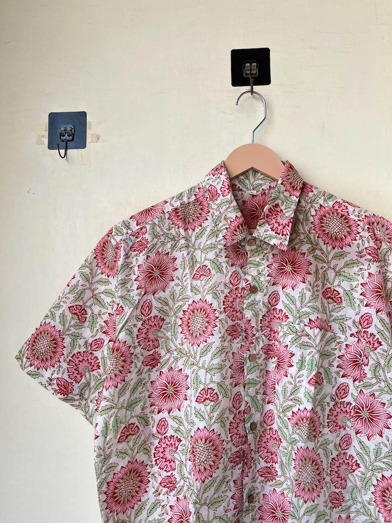 May include: Short-sleeved button-down shirt with a floral pattern. The shirt features a white background with a repeating pattern of pink flowers and green leaves. The shirt is hanging on a wooden hanger.