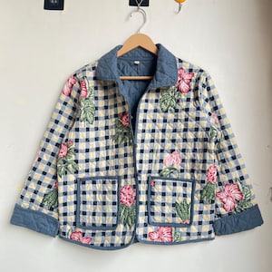May include: A quilted jacket in a blue and cream check pattern, decorated with pink floral embroidery. The jacket has a blue collar, cuffs, and pocket trim. It is suspended from a wooden hanger.